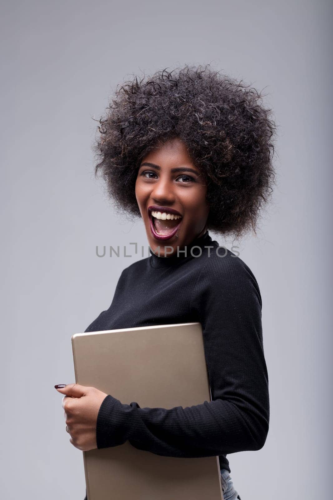 Young african american woman with an afro hairstyle is holding a laptop under her arm and smiling enthusiastically at the camera, expressing joy and excitement