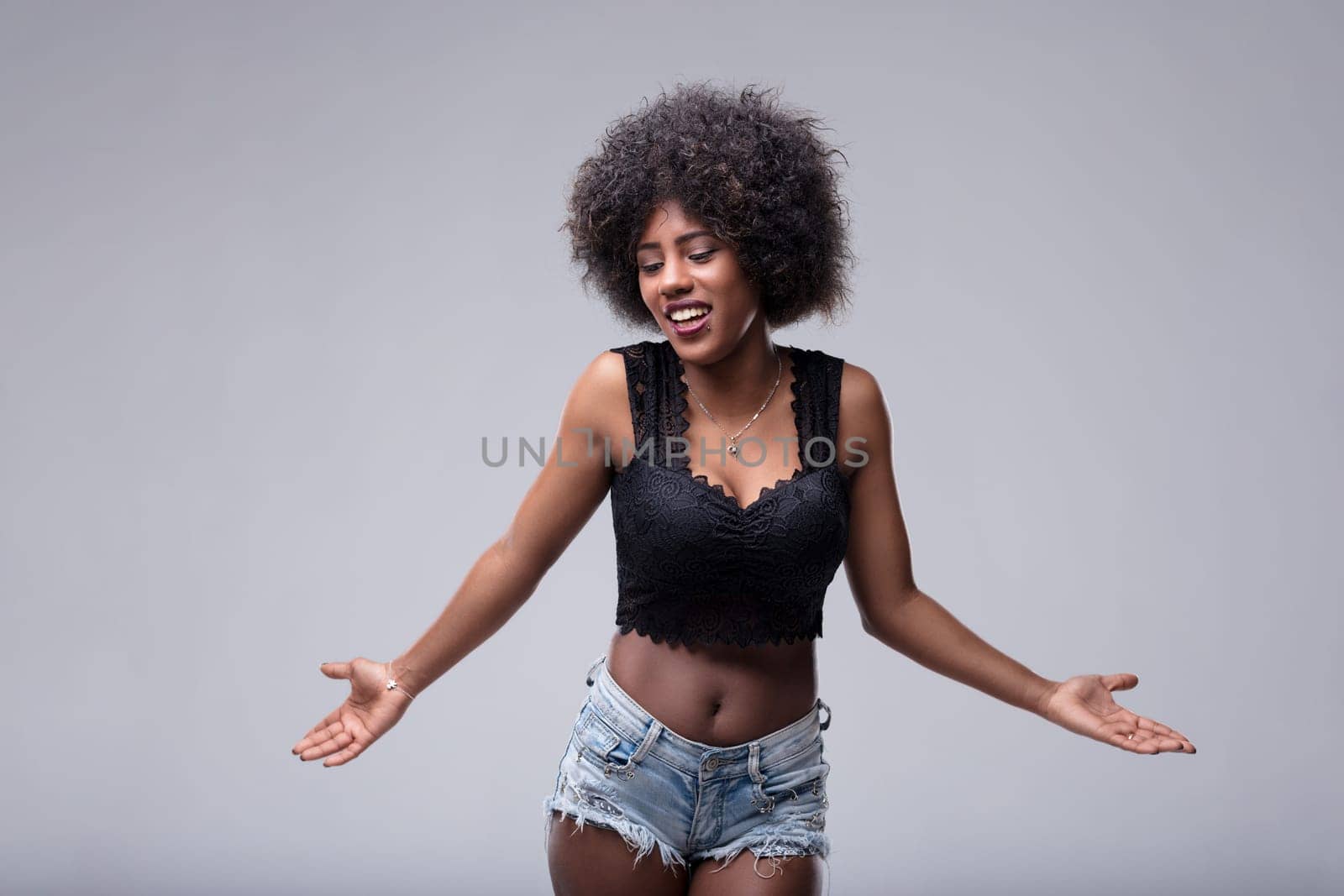 Young woman with an afro hairstyle is shrugging her shoulders with open arms and a confused expression, looking down and to the side against a gray background