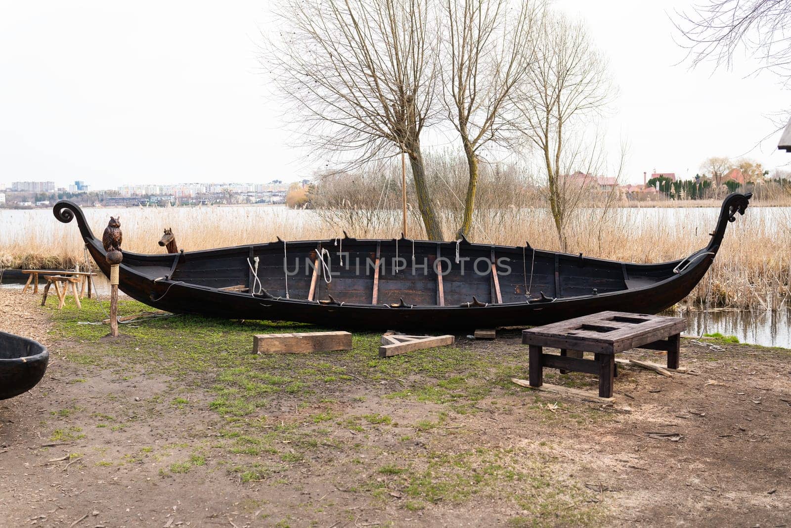 A traditional Viking boat on a grassy shore near the water, with wooden benches nearby and reeds in the background.