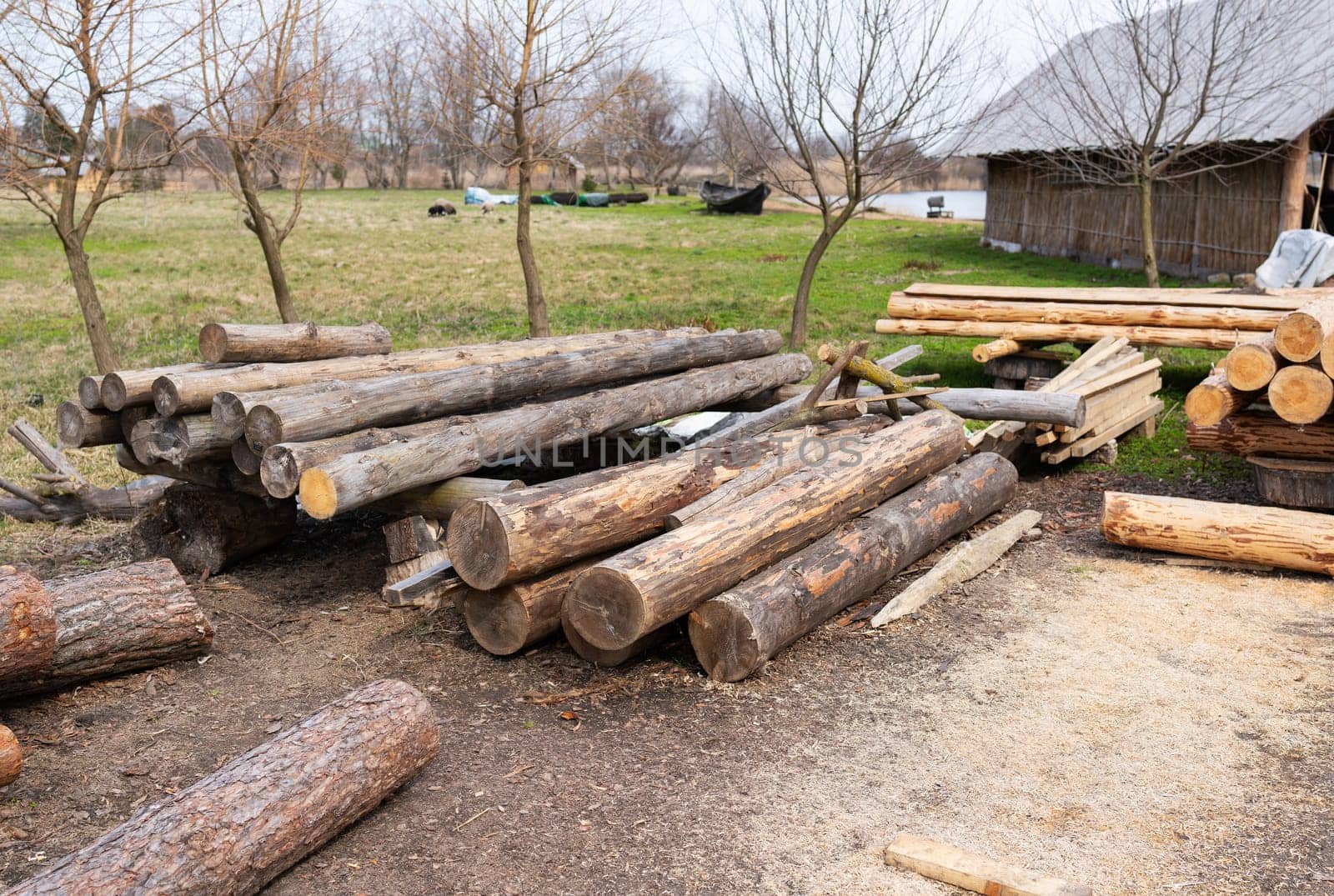 Stacked timber logs arranged in a rural area, surrounded by bare trees and grass, with a nearby barn in the background.