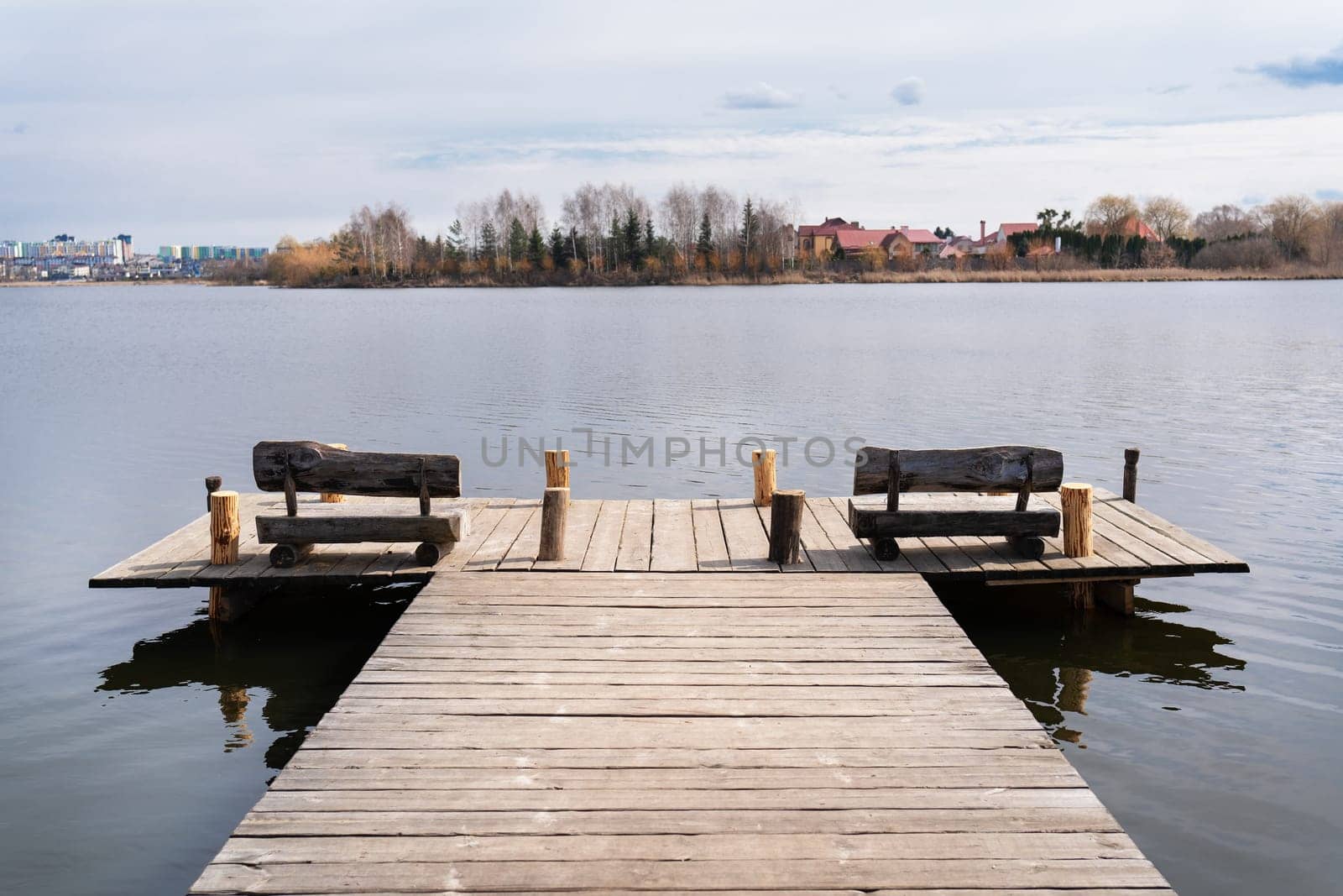 A wooden dock with rustic benches extends over a calm lake, with trees and houses visible in the distance.