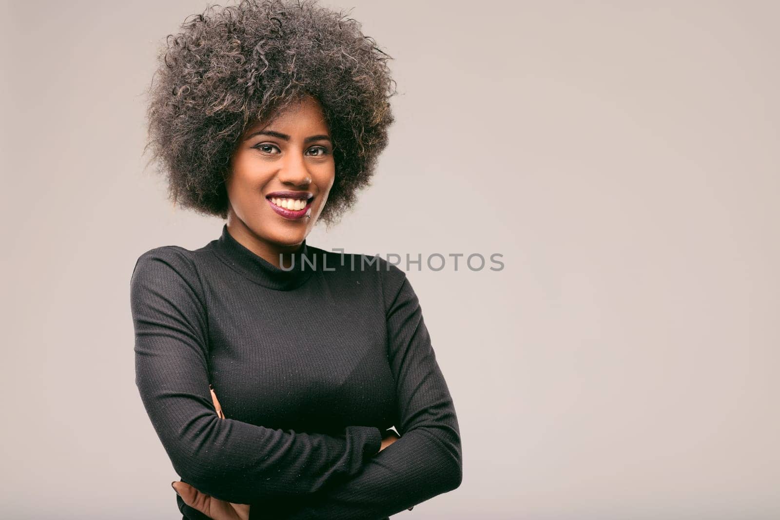 Studio portrait of a young smiling confident businesswoman with folded arms and afro hairstyle, wearing black turtleneck, looking at the camera