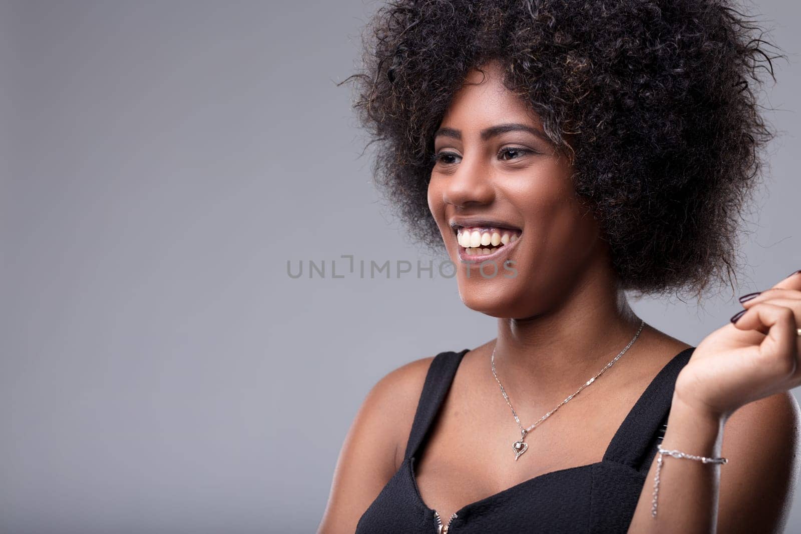 Studio portrait featuring a cheerful young woman with beautiful afro hair, smiling brightly while looking away, radiating happiness and positive emotions in a stylish black dress
