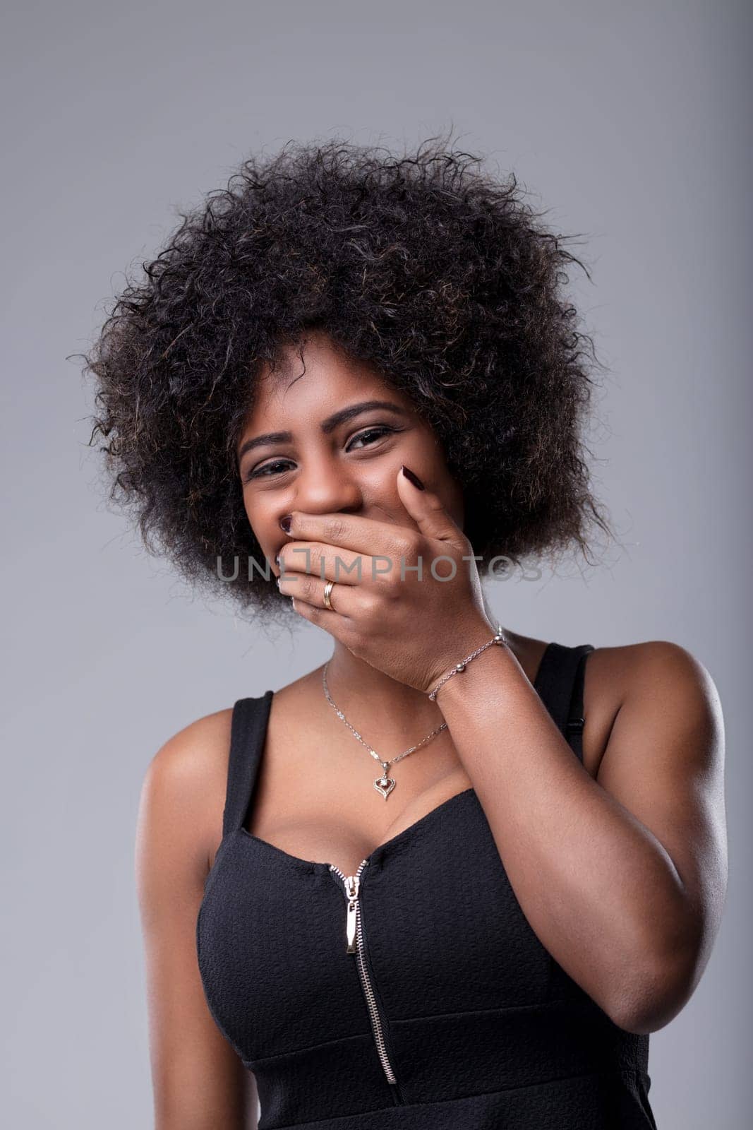 Studio portrait of a cheerful young woman with curly hair, wearing a black top, covering her mouth with her hand while laughing against a gray background