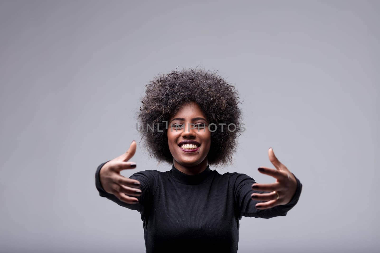 Studio shot of a young woman with afro hair, wearing a black turtleneck, opening her arms for a hug, smiling and looking at the camera, expressing joy and affection