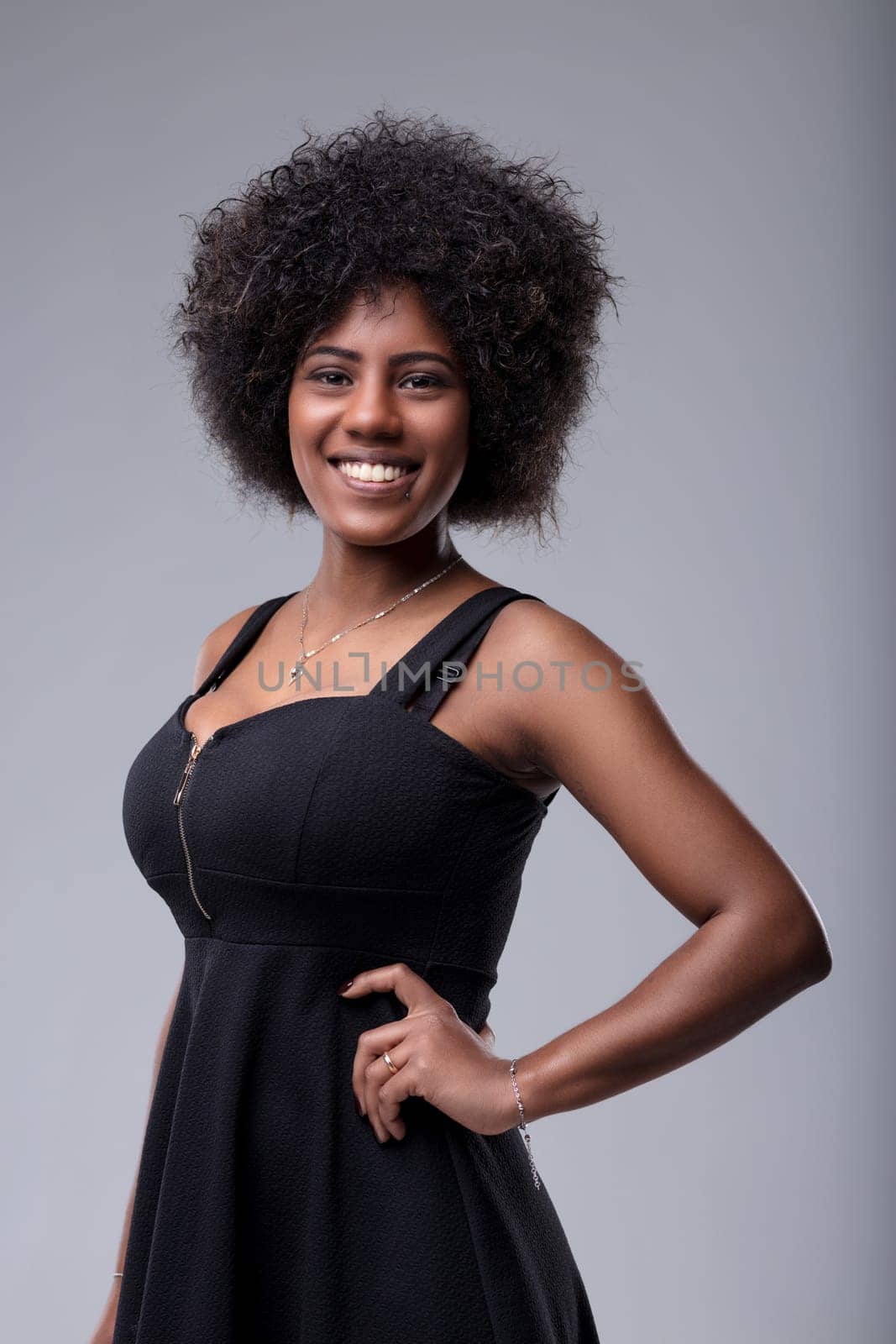Studio portrait of a cheerful young black woman with curly hair, wearing a black dress, smiling confidently while posing with her hand on her hip against a gray background