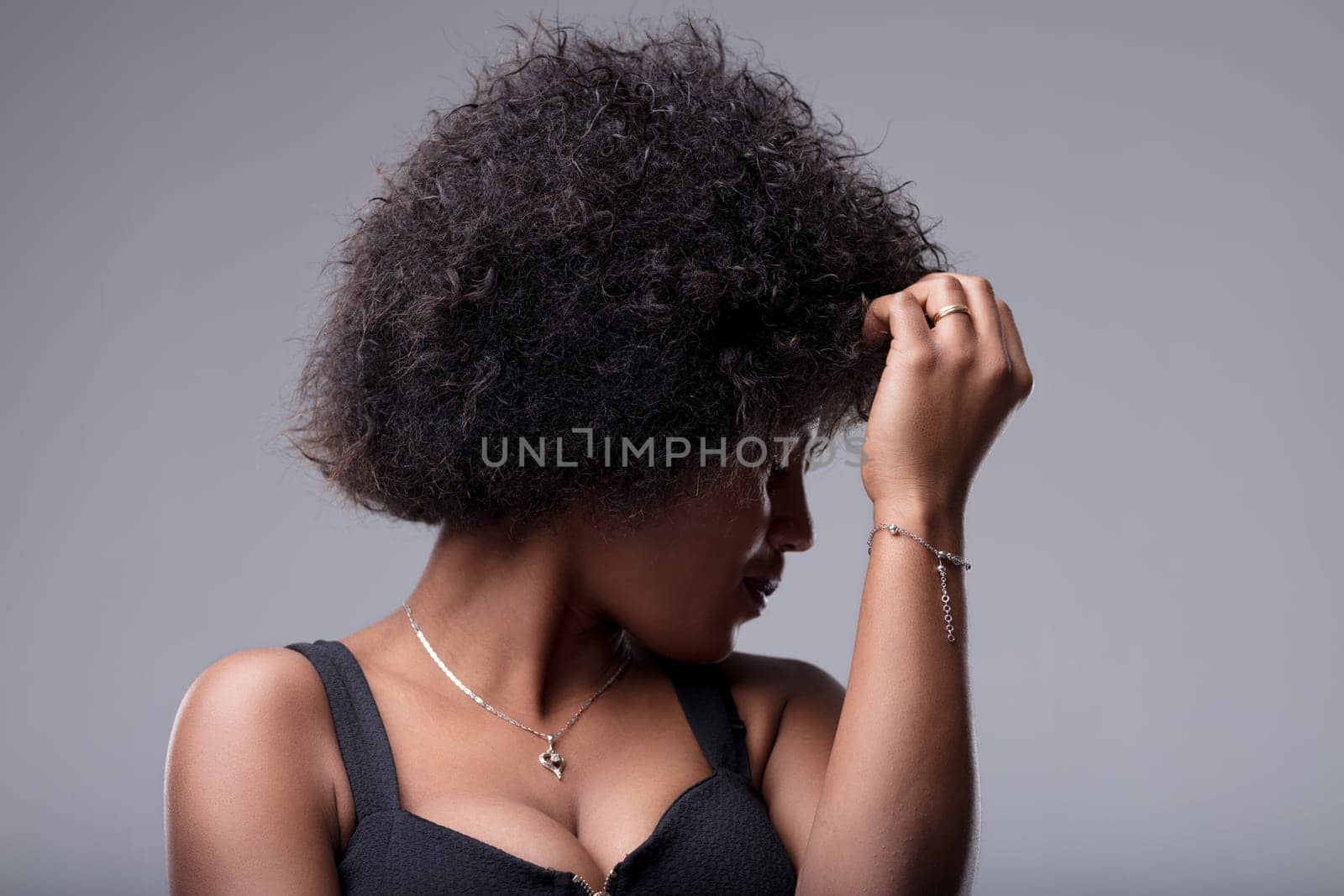 Studio portrait of a young black woman touching her afro hair while looking down, expressing shyness and introspection on a gray background