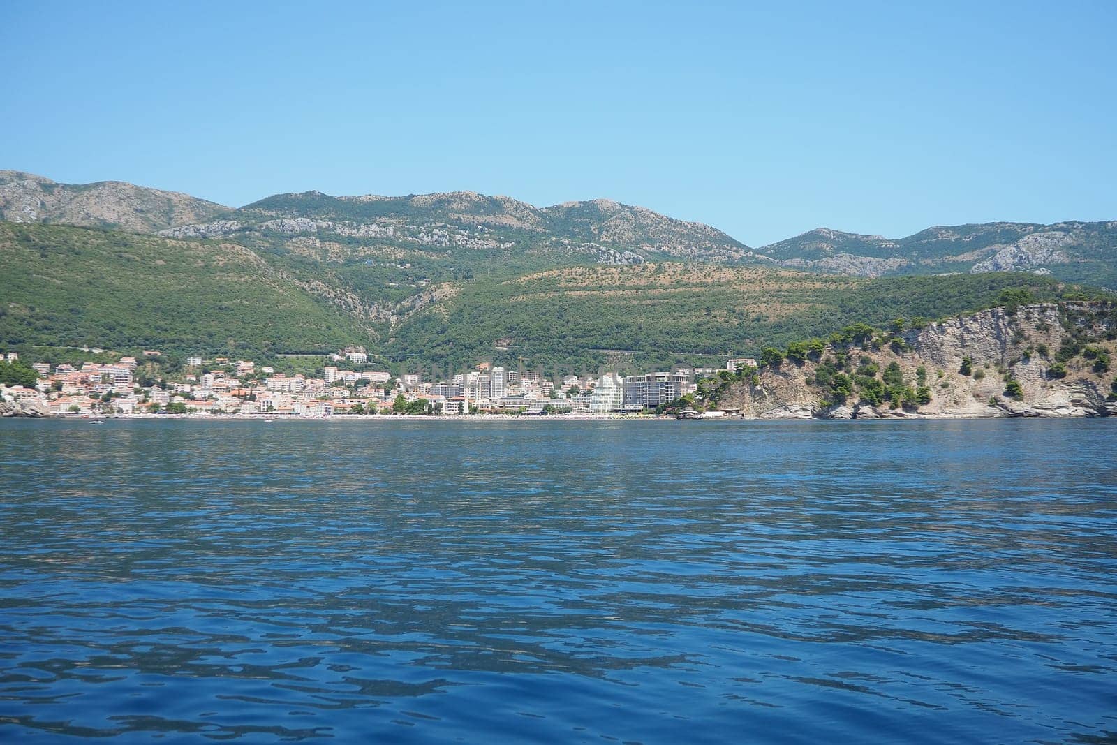Petrovac, Montenegro 07.22.24 A boat ride past shore with vacationers, sunbathers and swimmers. Hotels, sun umbrellas, Adriatic waves, buoys. Beach tourism. Vacation travel. Summer day. Sandy pebble.