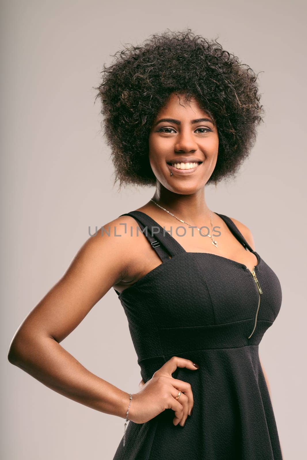 Studio portrait of a cheerful young latin american woman with curly hair, wearing a black dress, smiling broadly and posing with her hand on her hip against a light gray background