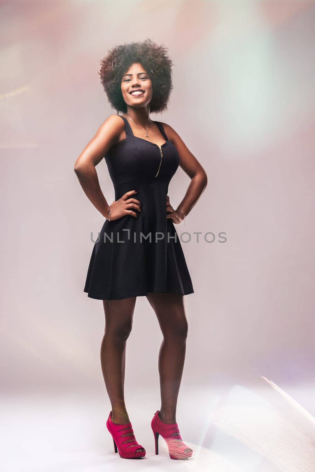 Full-length studio portrait featuring a fashionable young woman with an afro hairstyle, wearing a short black dress and pink high heels, confidently smiling at the camera