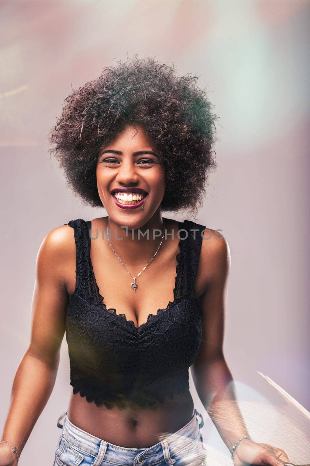 Portrait of a cheerful young woman with curly afro hair, beaming with a big smile while wearing a stylish black top and jeans, engaging directly with the camera
