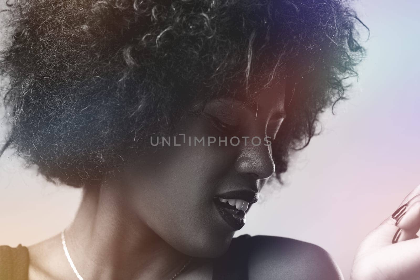 Black and white portrait featuring a young woman with an afro hairstyle, posing in a studio illuminated by colorful light effects, looking down and radiating a joyful smile