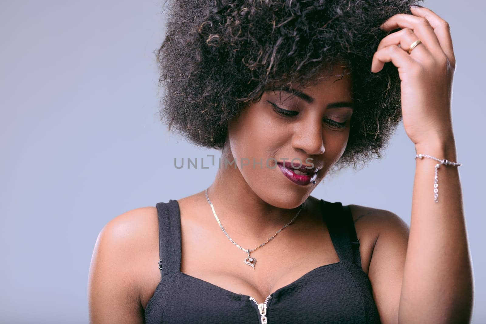 Young latin american woman with curly afro hair and dark skin is touching her hair while looking down with a slight smile on her face, expressing feelings of shyness and femininity