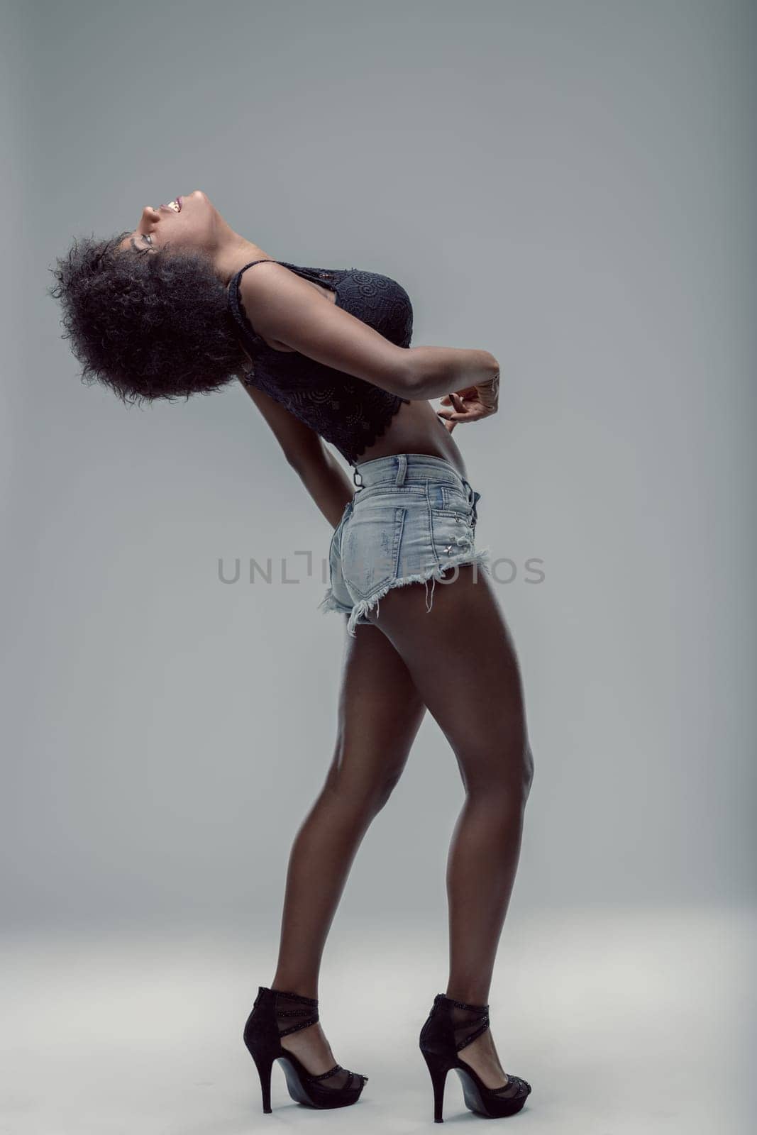 Young latin american woman with curly hair, wearing denim shorts and a black top, is posing in a studio, bending her back and looking up, expressing confidence and sensuality