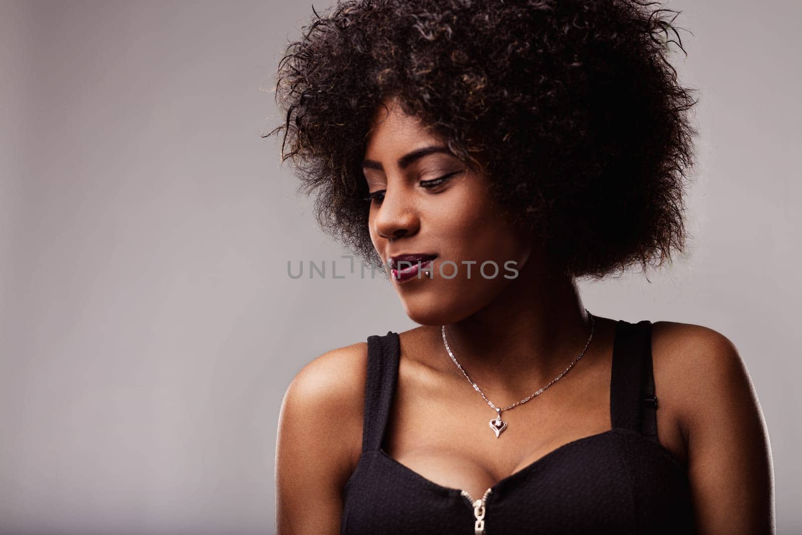 Studio portrait featuring a stylish young woman with an afro hairstyle, dressed in a black top and necklace, looking down with a subtle smile, exuding confidence and elegance