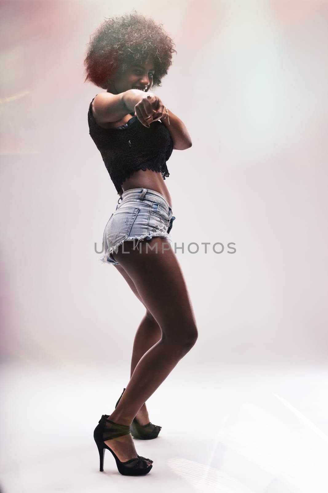 Young woman dancer with vibrant afro hair, wearing stylish denim shorts and high heels, smiling brightly while pointing directly at the camera, exuding confidence and joy