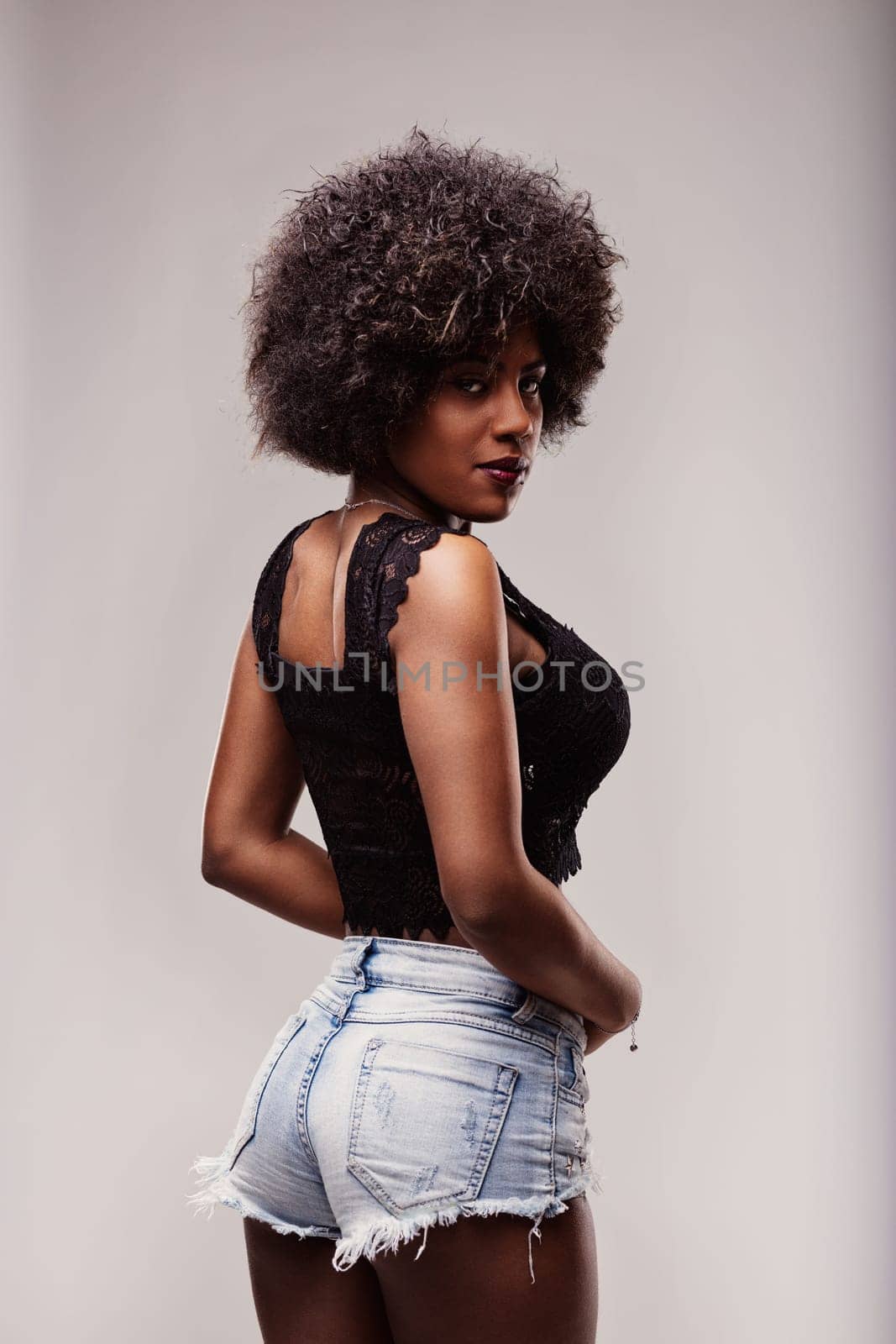 Studio portrait featuring a fashionable young black woman with a striking afro hairstyle, wearing trendy denim shorts and a sleek black top while looking over her shoulder with confidence