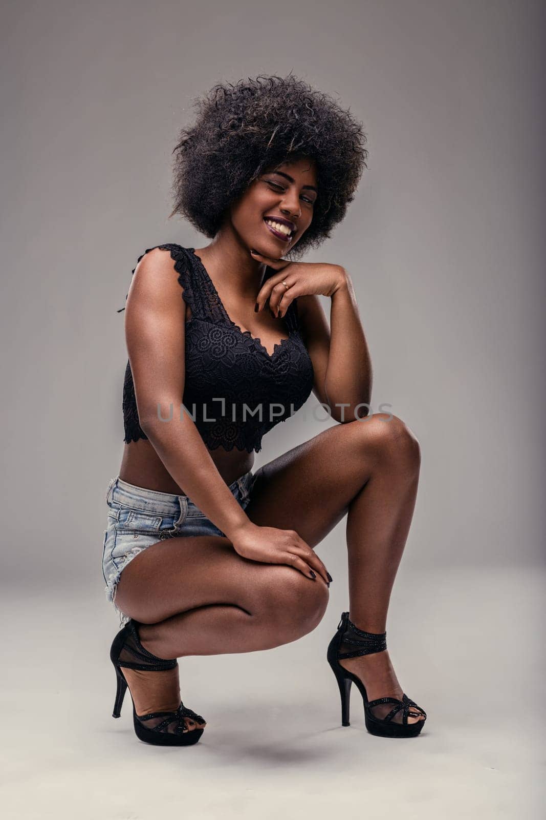 Studio shot featuring a beautiful young black woman with an afro hairstyle, crouching and smiling at the camera while wearing stylish denim shorts and a sleek black top
