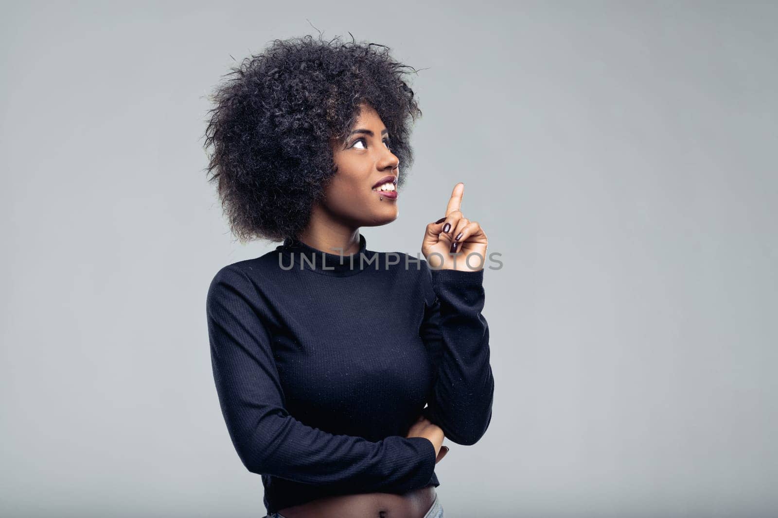 Young woman with vibrant afro hair looking upward and pointing, expressing a moment of inspiration and creativity, isolated against a smooth gray background