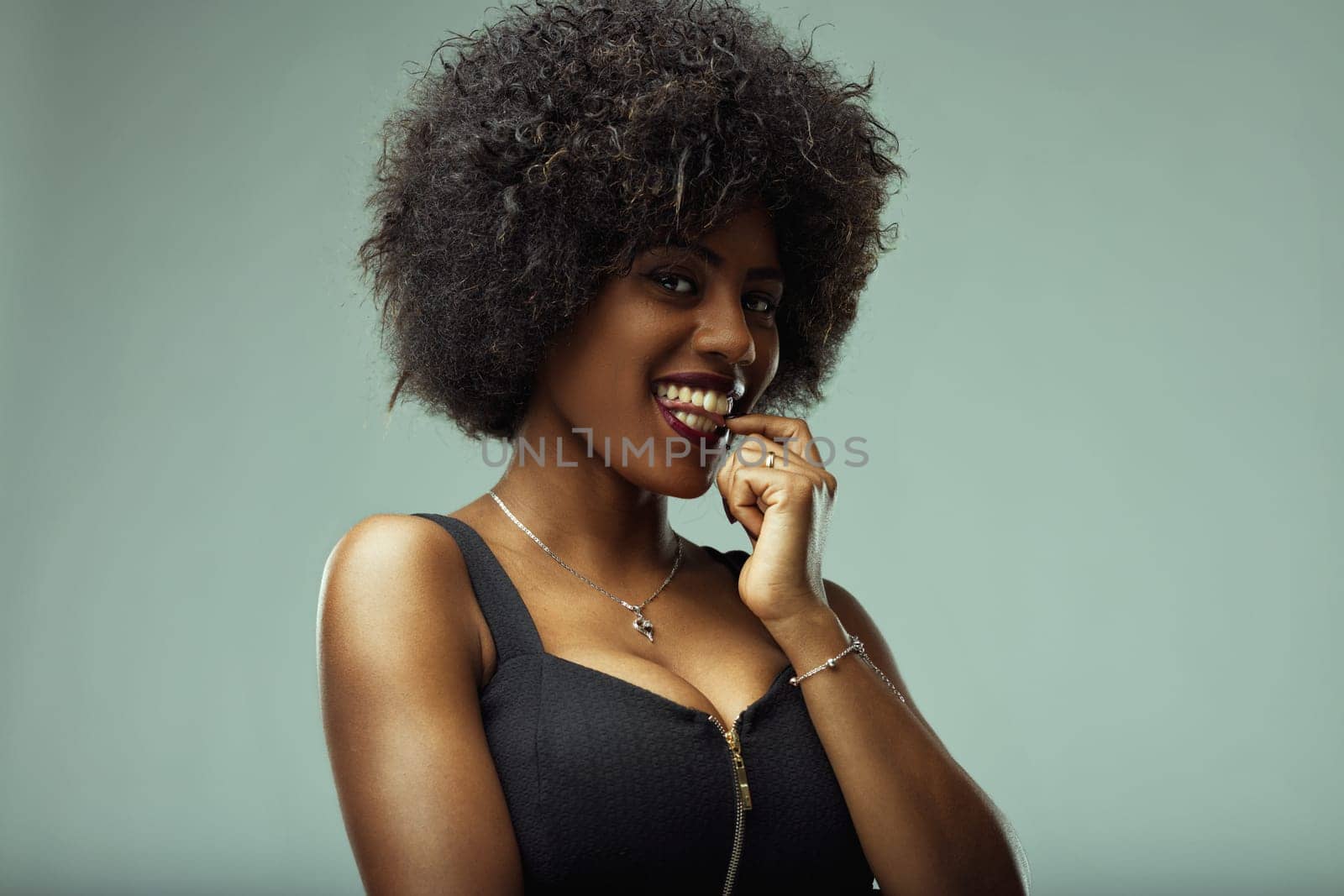 Studio portrait featuring a beautiful young latin american woman with curly hair, biting her fingernail and smiling joyfully, radiating positive emotions and charm