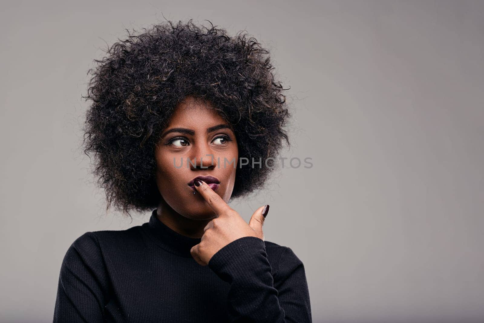 Studio portrait of a thoughtful young black woman with afro hair, wearing a black turtleneck, touching her lips with her finger while looking away