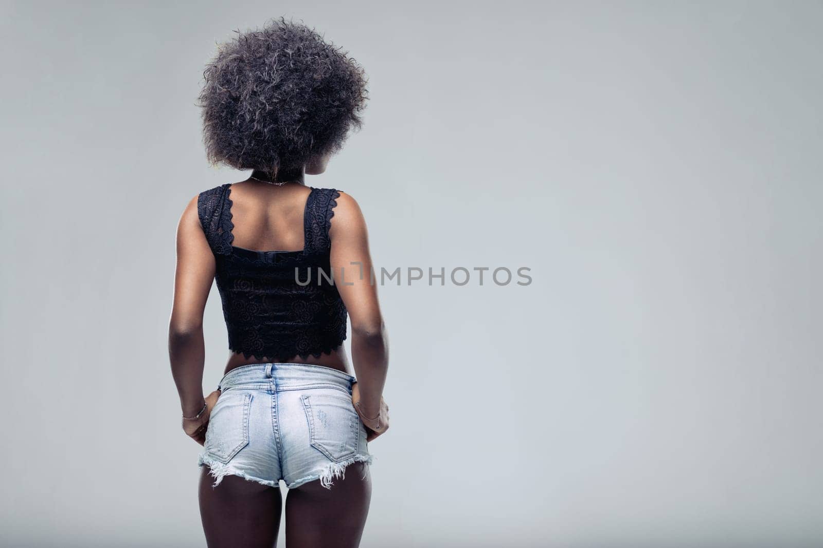 Rear view of a young black woman with an afro hairstyle, confidently posing with hands on her hips. Sporting denim shorts against a stylish gray background, exuding modern elegance