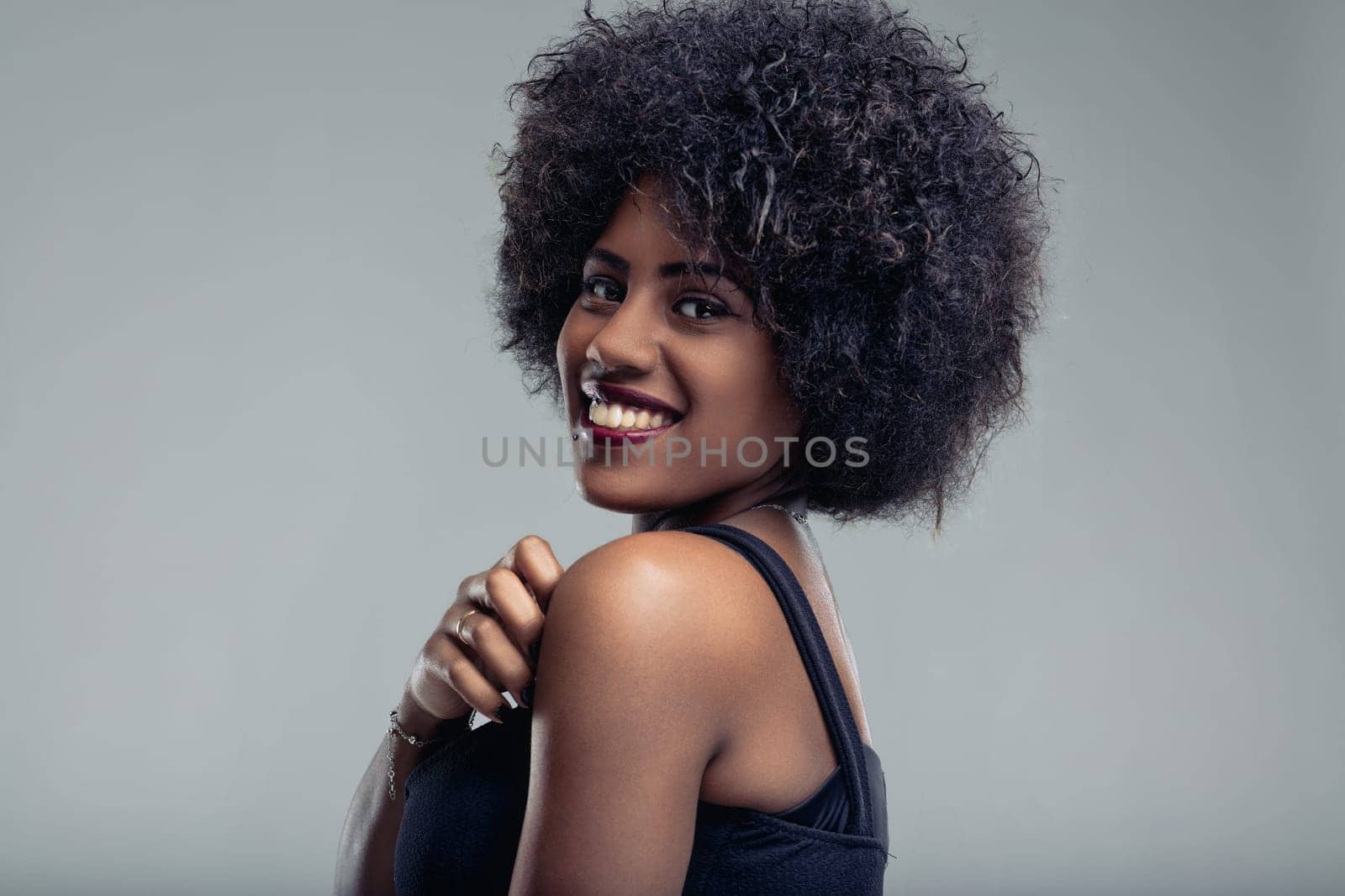 Studio portrait showcasing a beautiful young black woman with a curly afro hairstyle, smiling joyfully over her shoulder against a soft gray background, radiating confidence and happiness