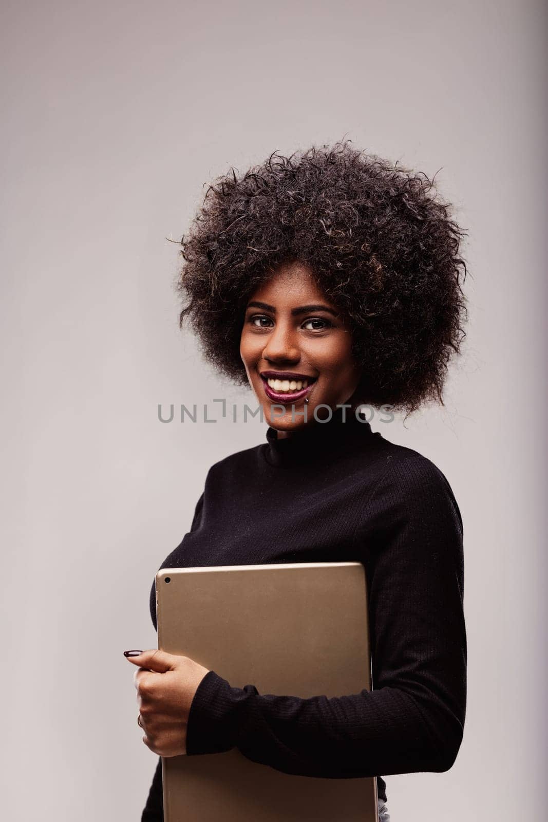 Smiling young black woman holding a tablet computer, exuding confidence and positivity, poses against a clean, gray studio background, showcasing her modern, professional style