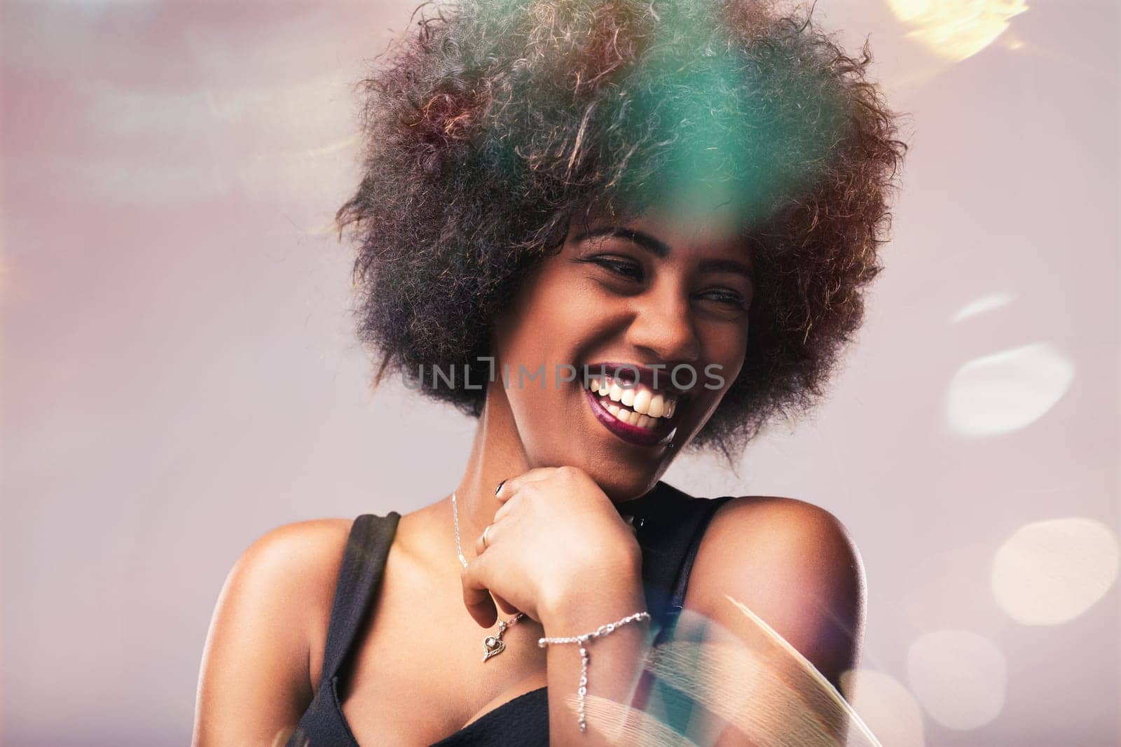 Cheerful young woman with an afro hairstyle smiling while touching her chin and looking away, radiating joy and confidence in a stylish tank top, necklace, and bracelet