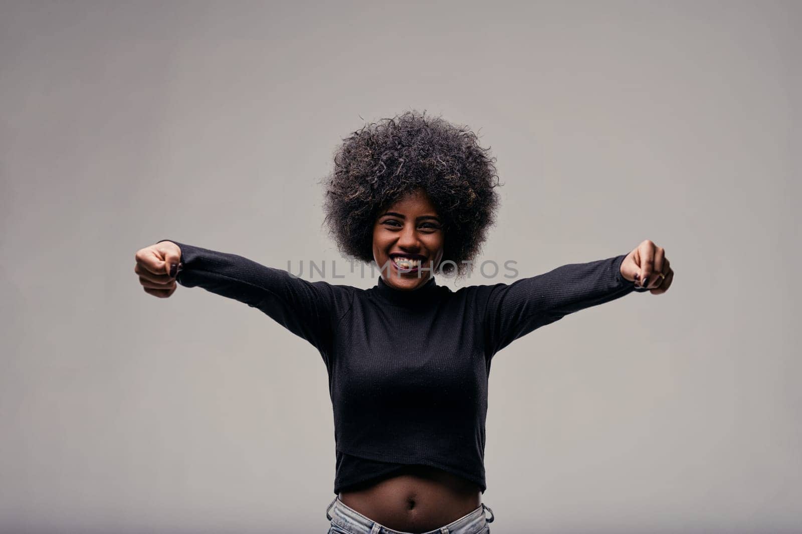 Cheerful young african american woman with big curly hair, wearing a black turtleneck, smiling and stretching her arms forward with clenched fists, radiating strength and happiness
