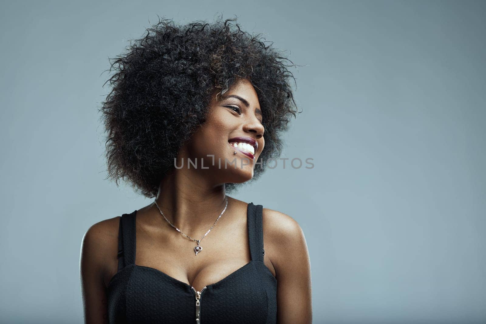 Studio portrait of a cheerful young latin american woman with curly hair, wearing a black top and a necklace, smiling and looking away against a gray background