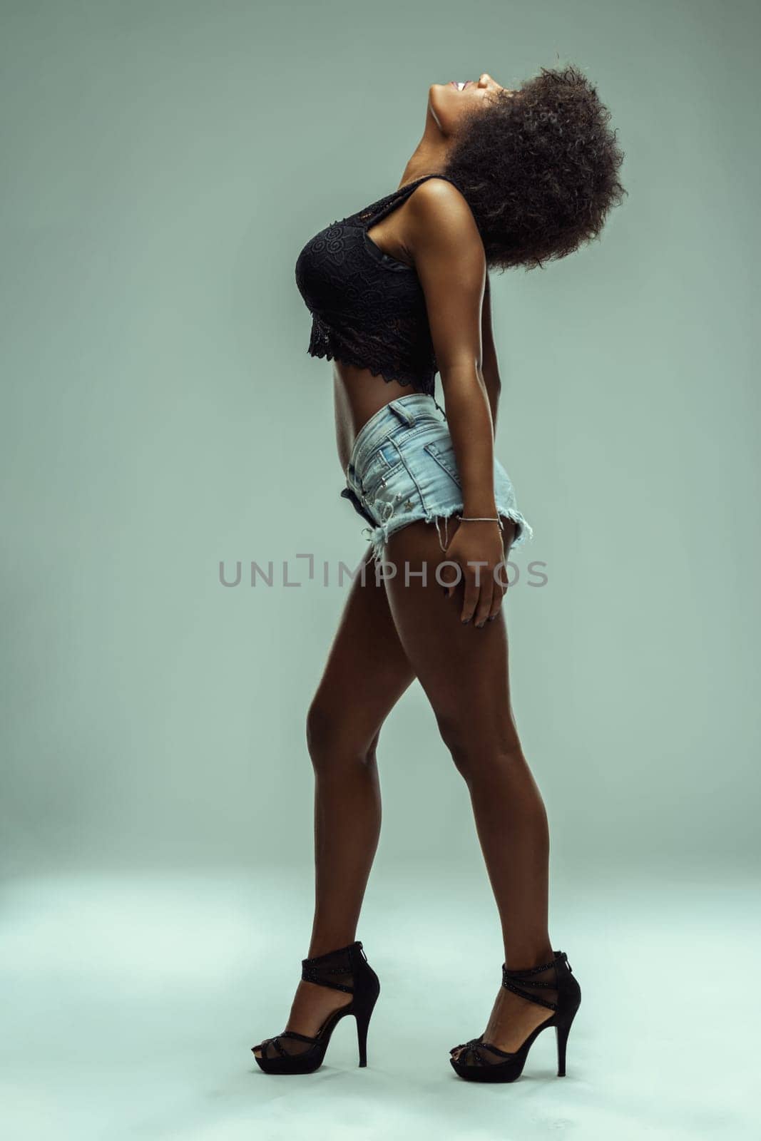 Young latin american model with afro hairstyle posing in a studio, wearing denim shorts, black top and high heels, expressing freedom and confidence