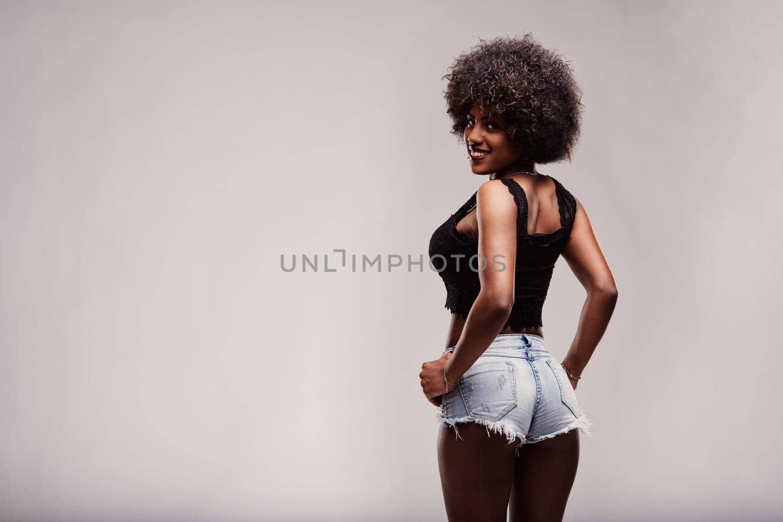 Studio shot of a beautiful smiling latin woman with afro hair, wearing denim shorts and black top, looking back over her shoulder with hands on hips, feeling confident and free