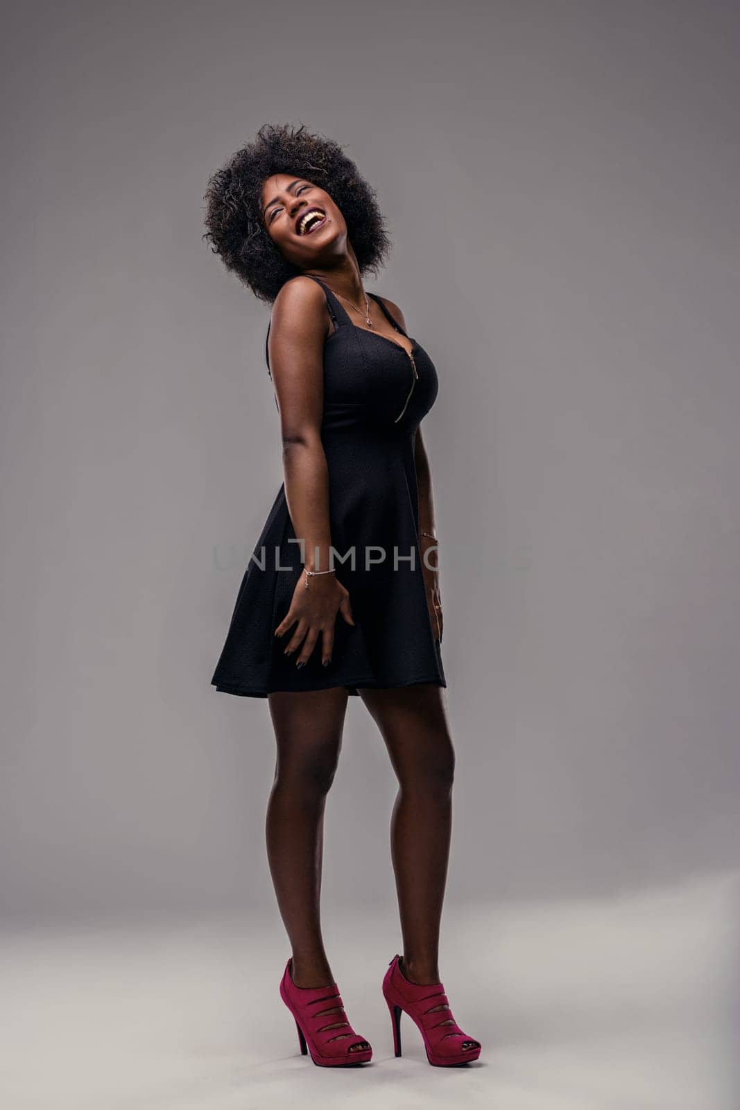 Studio shot of a cheerful latin american woman with afro hair wearing a black dress and red high heels, laughing and enjoying herself, expressing freedom and confidence