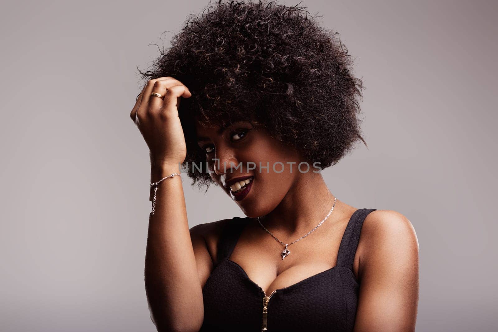 Studio portrait featuring a beautiful young latin american woman smiling while touching her curly afro hair, exuding confidence and sensuality against a soft gray background