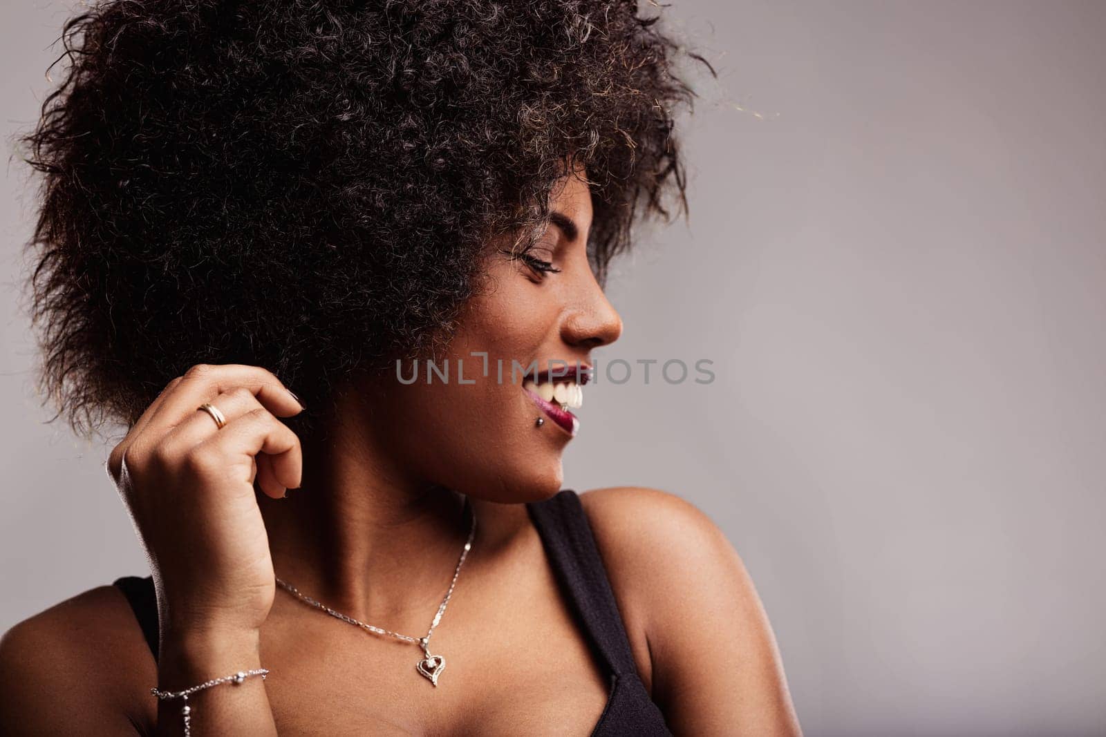 Smiling latin american woman with curly hair, touching her hair and gazing away, radiating freedom and confidence in a stylish studio portrait against a gray background