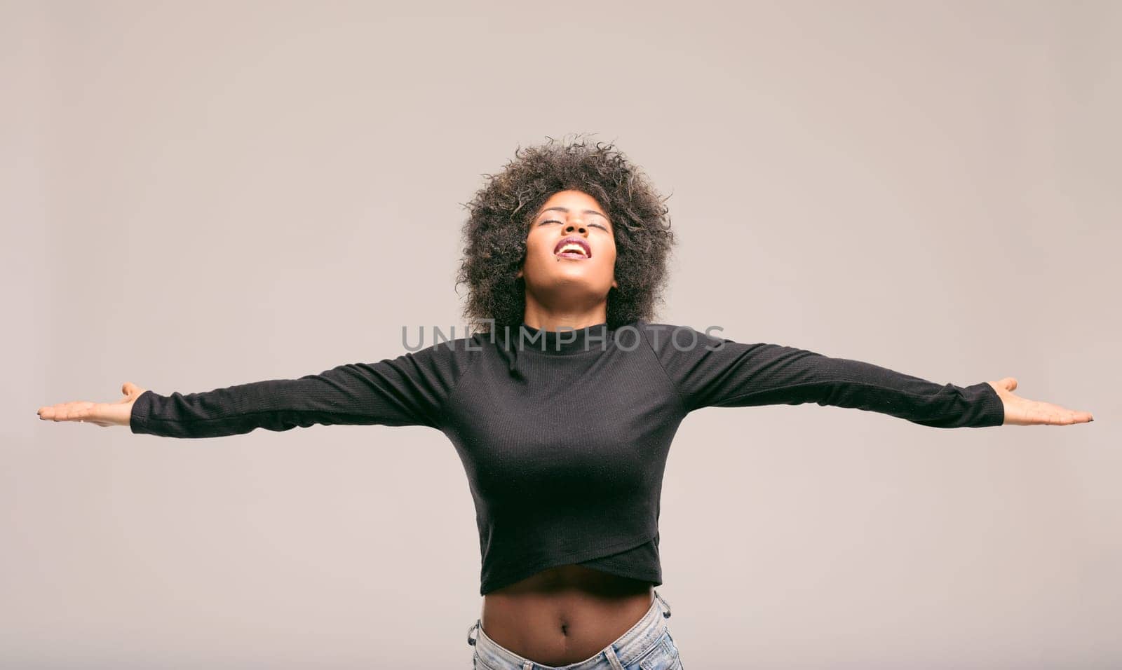 Young latin american woman with curly hair, wearing a black long-sleeved shirt and jeans, is expressing freedom and happiness with open arms and closed eyes, feeling like a diva