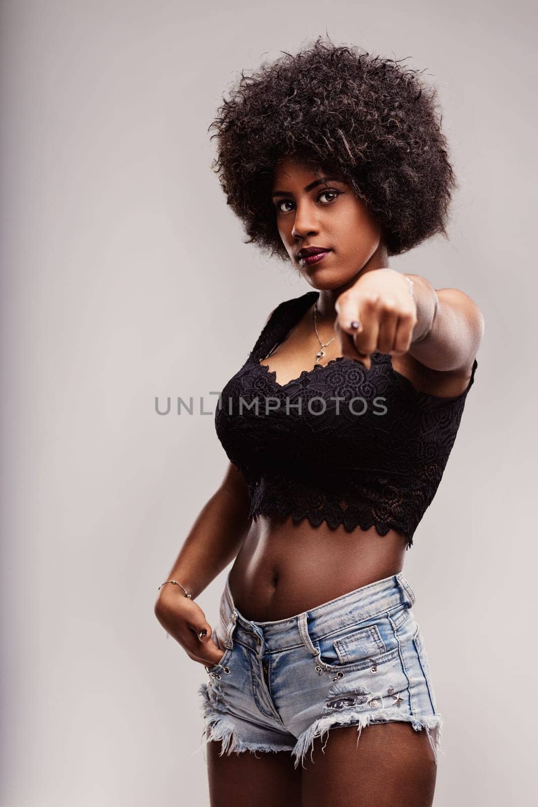 Young latin woman with afro hairstyle pointing her finger while posing confidently in a studio setting, wearing a black top and denim shorts, exuding a sense of empowerment and freedom