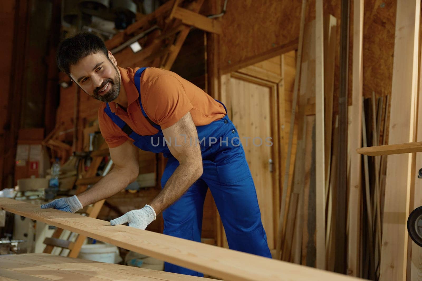 Young man carpenter in uniform putting wood plank on modern equipment closeup view. Experienced workman enjoying DIY woodworking project at his own small furniture factory