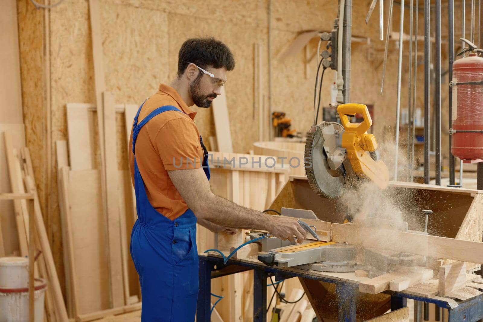 Skilled concentrated male carpenter wearing coveralls and protective glasses sawing wood plank operating with round circular saw at carpentry workbench