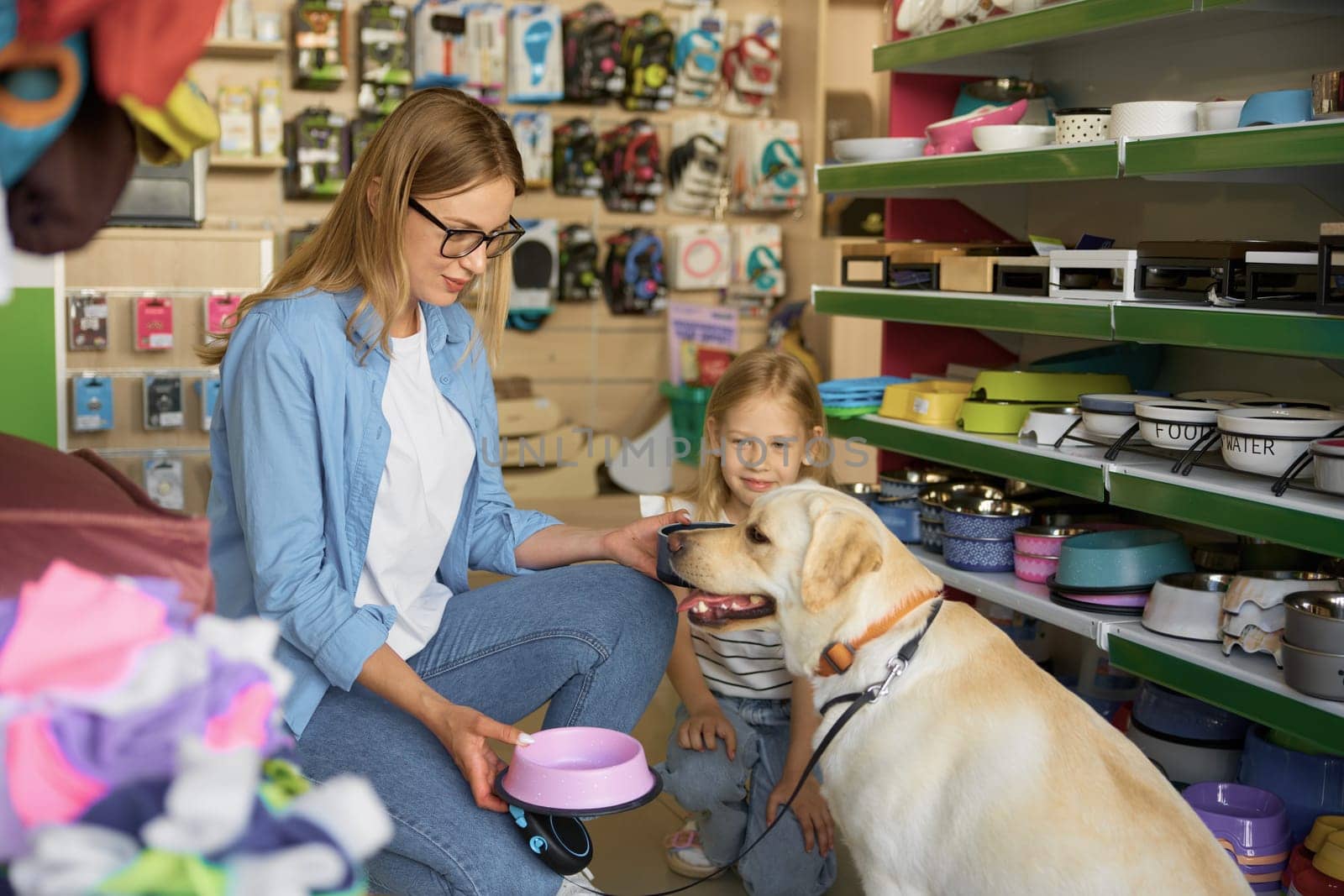 Mother and toddler daughter choosing new plastic food plate for Labrador dog enjoying shopping at pet store