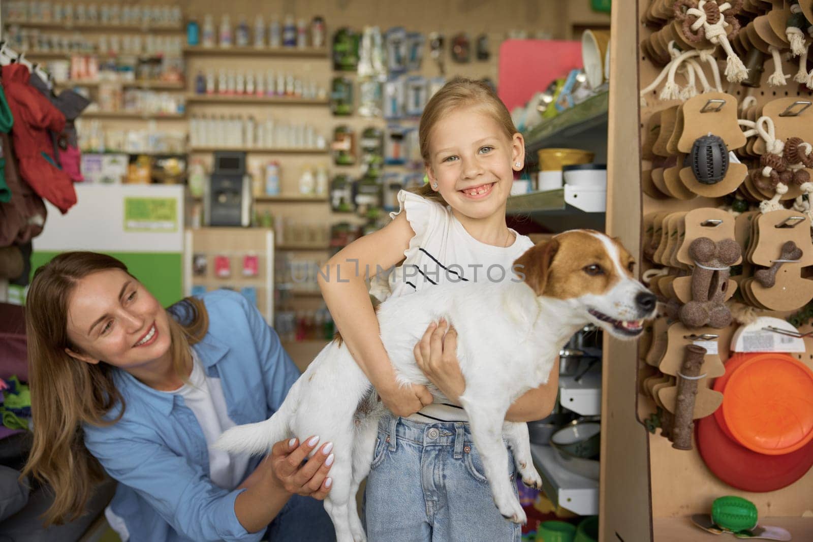 Mother helping cute little girl posing with dogs in hands over pet shop background. Happy time together and family shopping enjoyment