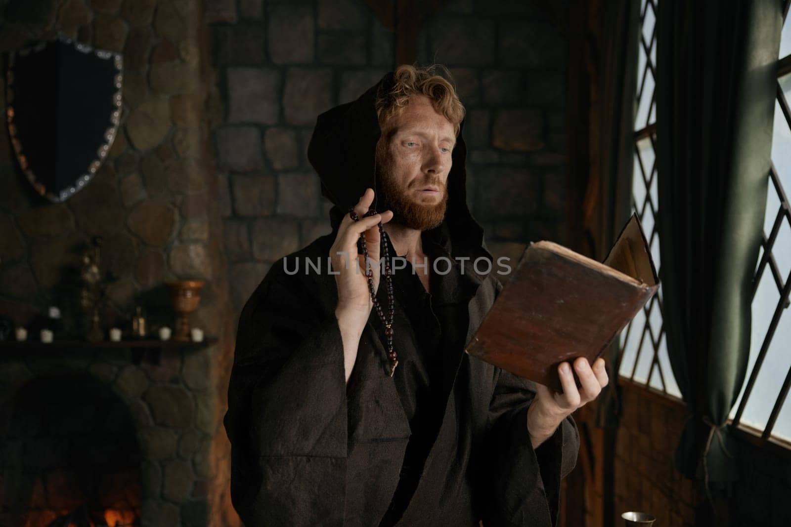 Medieval monk wearing robe with hood reading sacred religious book praying with rosary beads in hand