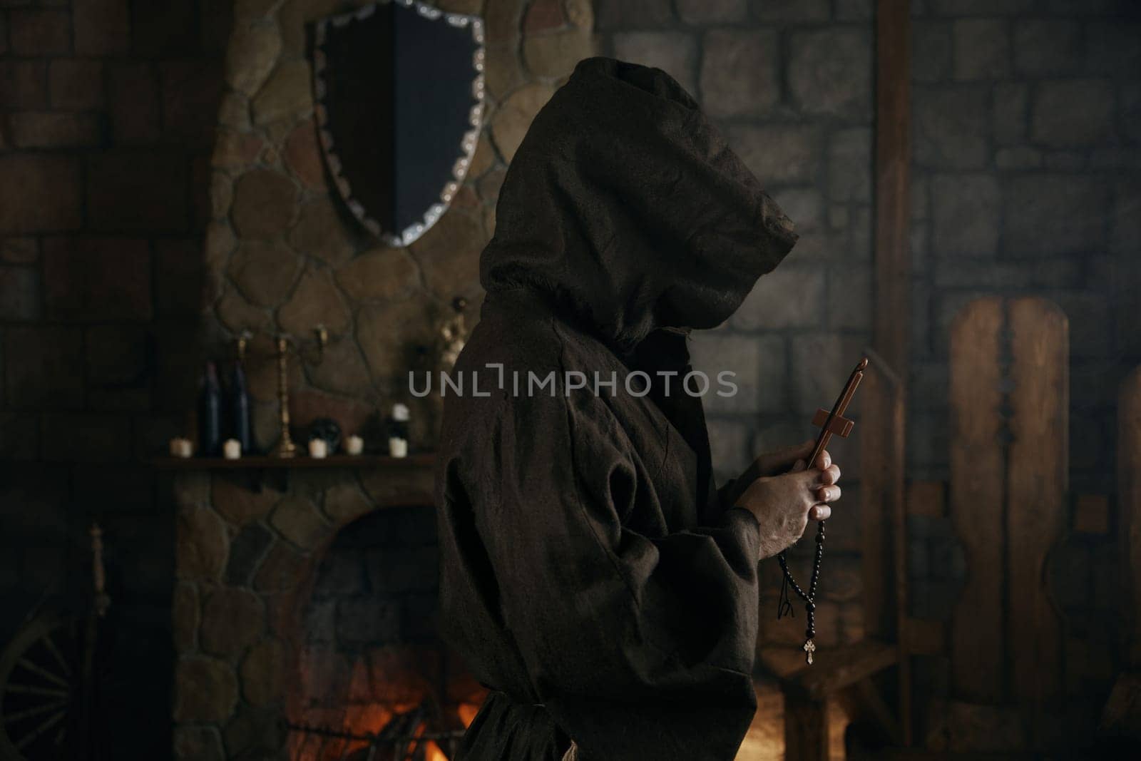 Faithful priest in robe hood praying with rosary beads and cross over ancient monastery interior. Side view portrait