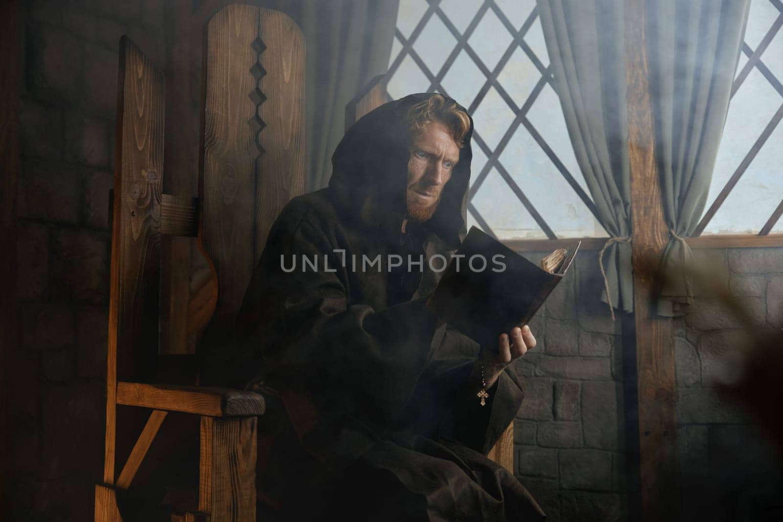 Old medieval monk wearing robe with hood reading sacred religious book praying with rosary beads in hand