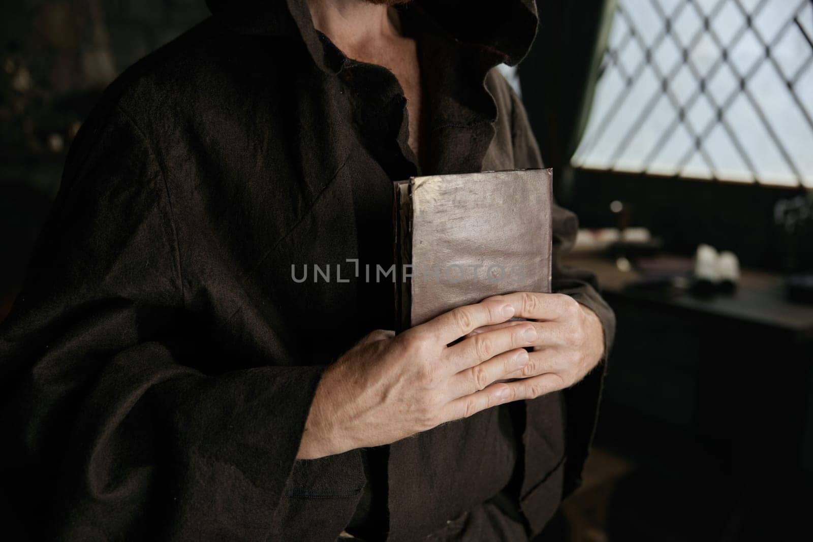Mature priest wearing medieval robe clothing holding closed bible book and rosary beads in hands closeup