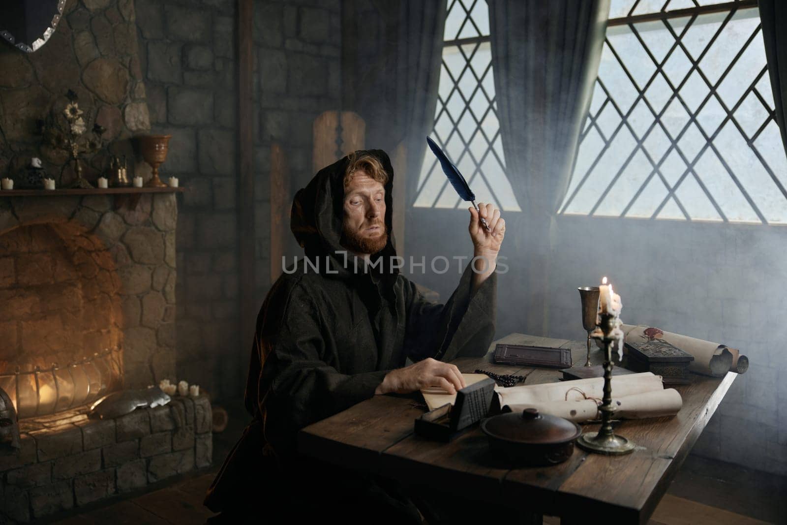 Medieval monk in cassock with quill pen writing historic manuscripts at table under candlelight in abbey cell