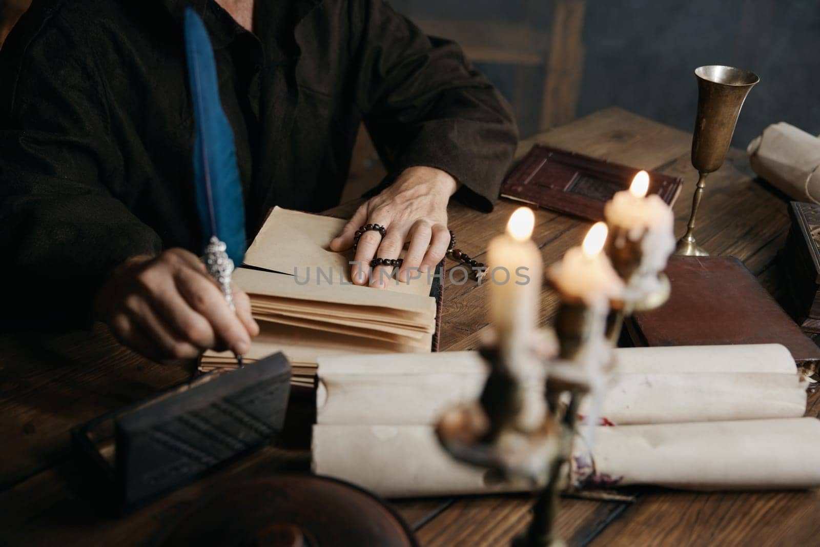 Medieval monk in cassock with quill pen writing historic manuscripts at table under candlelight in abbey cell