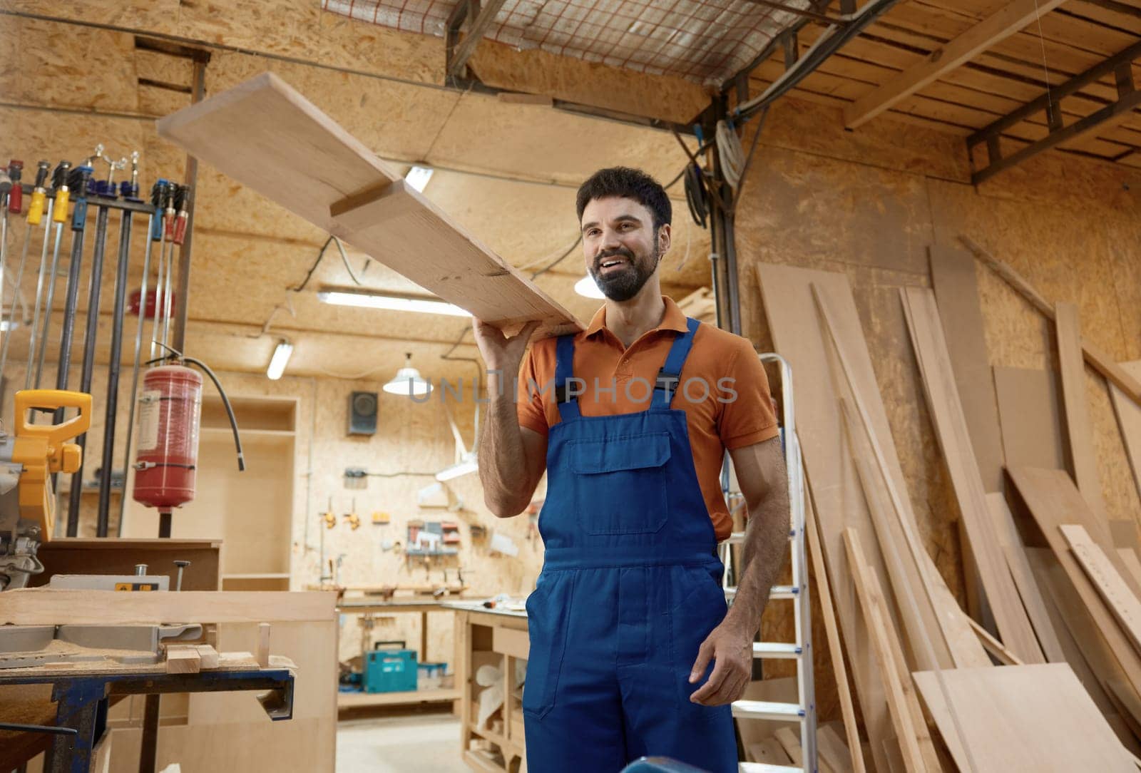 Smiling woodworker carrying timber plank on shoulder standing over carpentry workshop background. Woodworking and furniture manufacturing concept