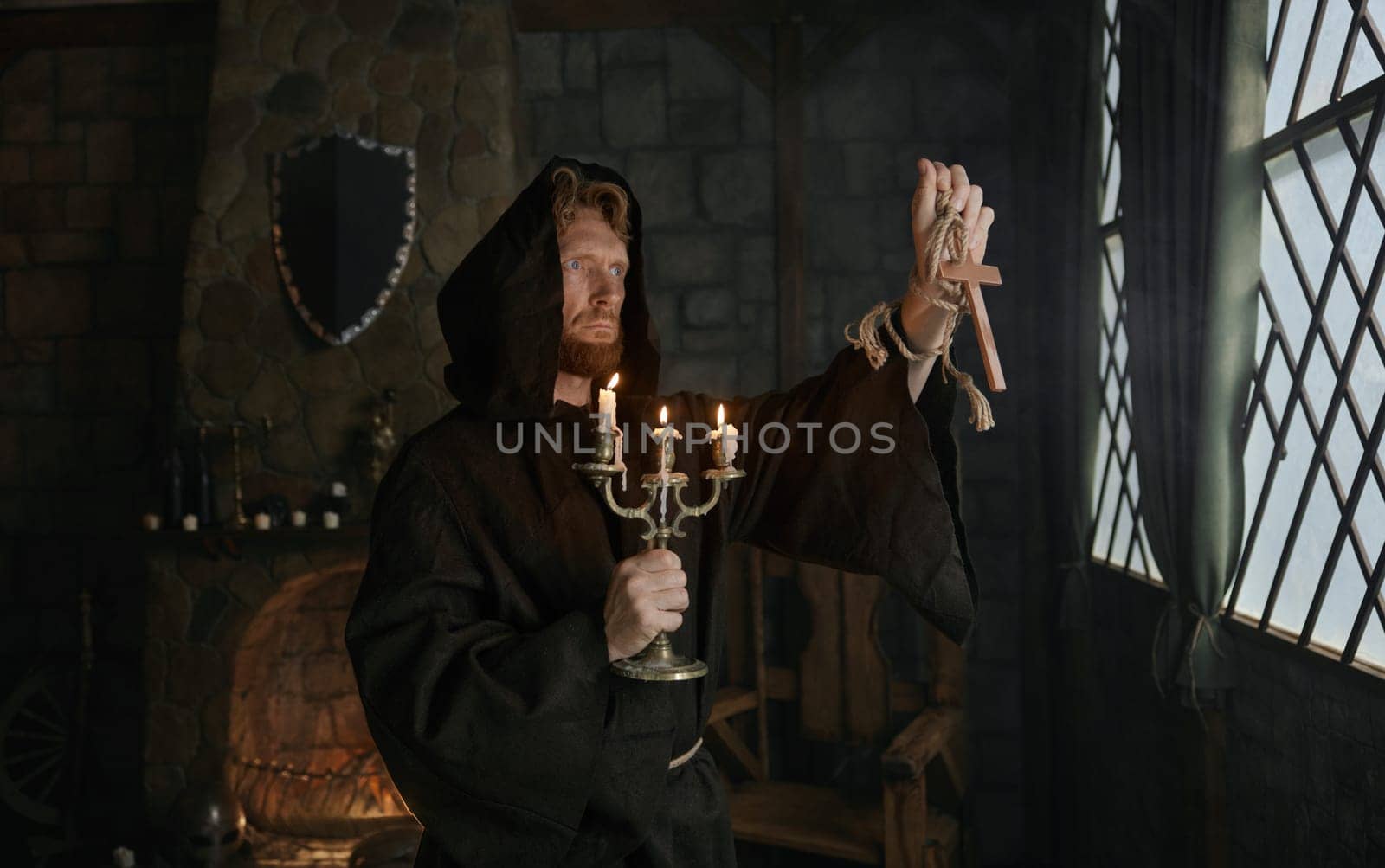 Portrait of hooded monk in medieval costume holding wooden cross and candlestick praying in church